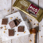 Porciones de turrón de chocolate con leche y cacahuete sobre papel, con cacahuetes y miel, junto a su envase Picó