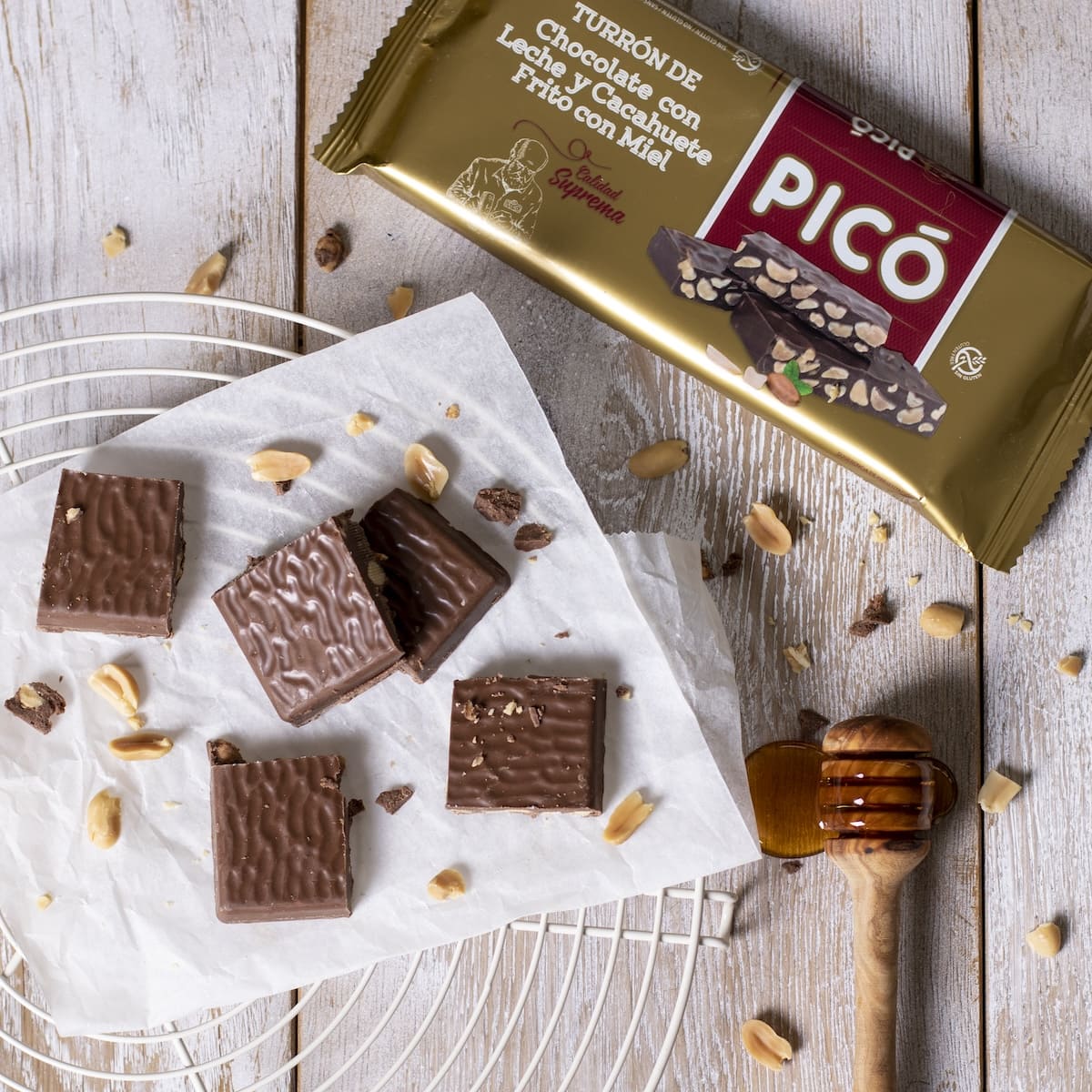 Porciones de turrón de chocolate con leche y cacahuete sobre papel, con cacahuetes y miel, junto a su envase Picó