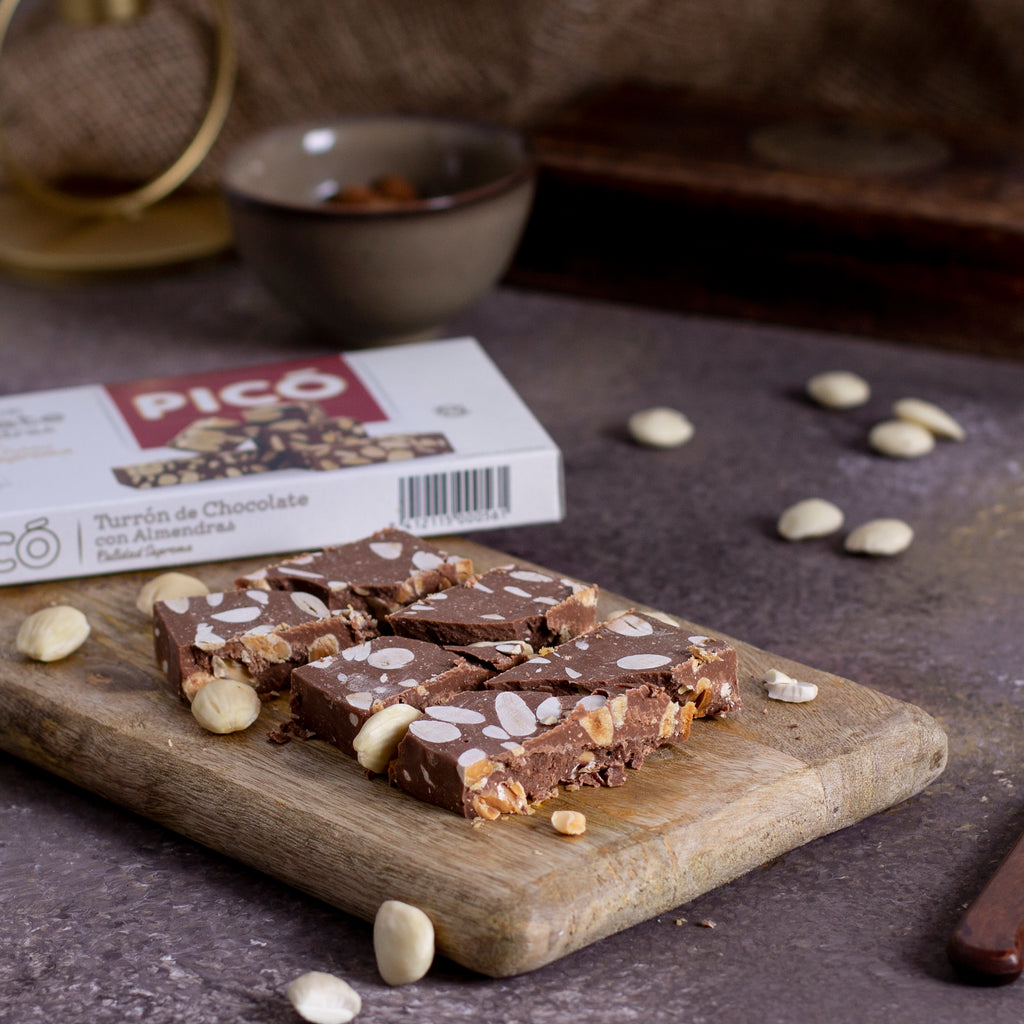 Turrón de chocolate con almendras cortado en dados sobre una tabla, con la caja Picó detrás