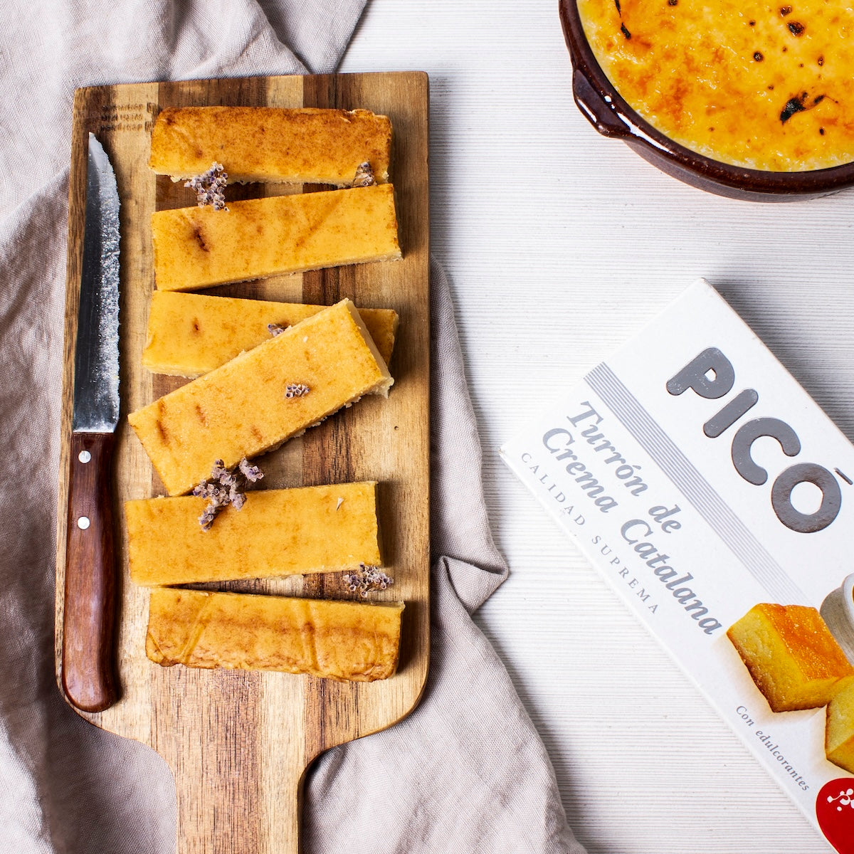 Turrón de crema catalana cortado en barritas sobre una tabla, junto a una crema catalana y su caja Picó